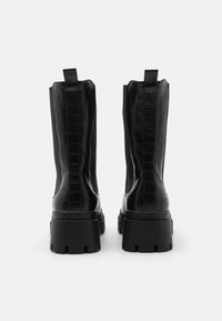 Bottines noires à motif cuir de crocodile, semelle plus épaisse et languettes à l'arrière pour un enfilage facile. Fini lisse et brillant.