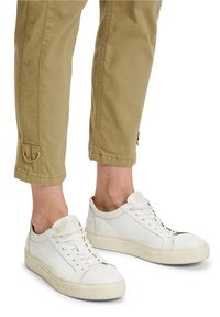 Bílé kožené tenisky s texturovaným povrchem, které mají ploché šňůrky, zaoblenou špičku a béžovou gumovou podrážku. Nošeny s khaki kalhotami do kotníků.