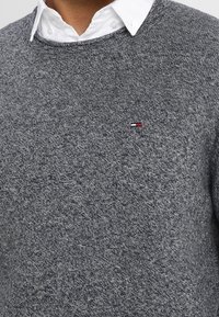 Harmaa neulepaita, jossa on pyöreä kaula-aukko ja pitkät hihat. Rintamuksessa on pieni punavalko-sininen logo.