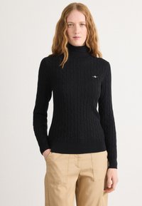 Mustra musta kabeli kudumütsiga turtleneck-särk, millel on soonikkoes varrukad ja alumine äär. Rinnal on brodeeritud logo. Kombineeritud khaki pükstega.