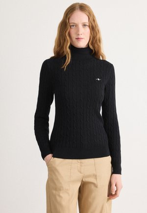 Black cable knit turtleneck sweater with ribbed cuffs and hem. Features embroidered logo on the chest. Paired with khaki trousers.