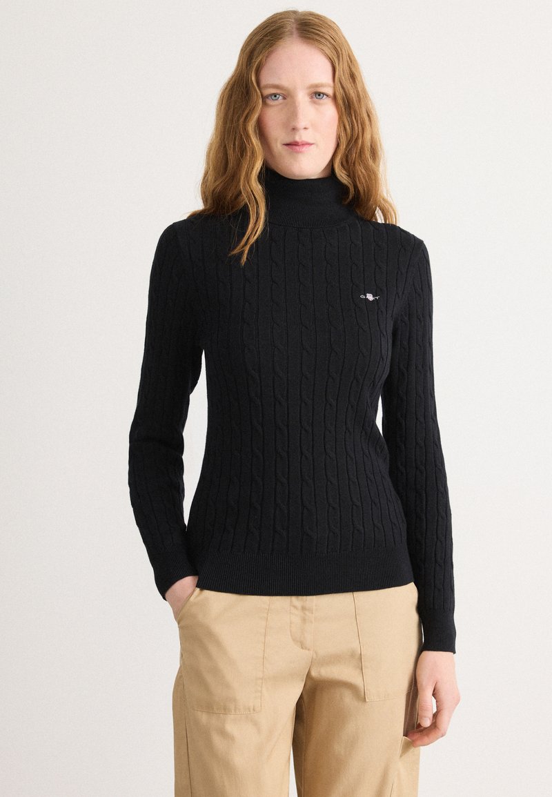 Mustra musta kabeli kudumütsiga turtleneck-särk, millel on soonikkoes varrukad ja alumine äär. Rinnal on brodeeritud logo. Kombineeritud khaki pükstega.