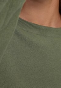 Pull léger couleur vert olive avec une texture douce, doté d'un col ras du cou et de poignets côtelés. Vue en gros plan mettant en avant les détails du tissu.