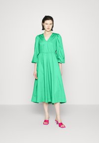 Vestido verde com decote em V, mangas volumosas, cintura marcada e saia fluida. Sapatos de salto alto abertos rosa. Tecido suave e brilhante.