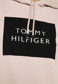 Lichtroze hoodie met een zwarte rechthoekige patch met daarop "TOMMY HILFIGER" in witte letters. Trekkoorden zitten aan de halslijn.