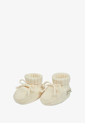 Bonnets de bébé tricotés en crème, avec un bord-côte à la cheville et un détail en nœud. Texture douce avec un bout arrondi et une petite étiquette sur le côté.