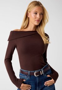 Top marrone scivolato sulle spalle in tessuto morbido, con maniche lunghe dotate di fori per il pollice. Abbinato a jeans di denim blu e una cintura marrone.