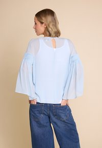 Lyseblå bluse med gjennomsiktige, utsvingte ermer og samlet detaljer. Nøkkelhullsåpning bak. Kledd med høyt liv denimjeans.