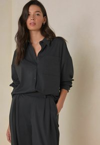 Long-sleeve black button-up shirt with a chest pocket, made from smooth fabric. Paired with matching black tailored trousers.