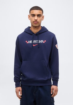 Mladý muž v tmavomodrej mikine s kapucňou s nápisom "HRVATSKA" a červeným logom Nike na prednej strane, stojaci pred svetlým jednofarebným pozadím.
