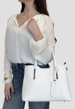 Donna con camicetta bianca e jeans che tiene una borsa bianca con etichetta, mostrando la mano con unghie rosse e braccialetto d'oro.