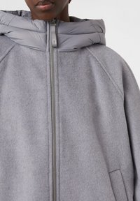 Person wearing a light gray zip-up coat with a padded hood and side pocket, shot from shoulders to chest.