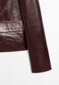 Burgunderfarbene Lederjacke mit glattem Textur, die einen subtilen Glanz und eine gestickte Taschenanzeige an der Seite aufweist.