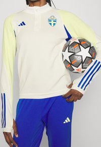 Maglietta bianca a maniche lunghe con accenti gialli e blu, con un crest della Svezia, abbinata a pantaloni blu e in possesso di un pallone da calcio bianco e nero.
