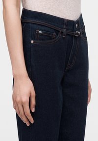 Gros plan d'une personne portant un jean taille haute bleu foncé avec des coutures orange et une petite poche avant étiquetée DKNY Jeans.