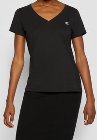 Černé tričko s krátkým rukávem a výstřihem do V vyrobené z hladké látky, se malým bílým vyšívaným logem na hrudi.