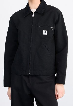 Osoba nosząca czarną kurtkę na zamek z kieszenią na piersi zapinaną na zamek i białą naszywką z logo, w połączeniu z czarnymi spodniami na jednolitym tle.