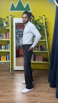 White ribbed turtleneck sweater, dark jeans with visible stitching, and white sneakers. Background features shelves with books and plants.