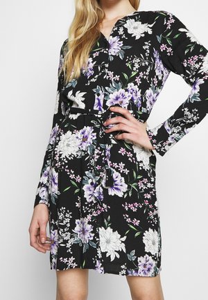 Kobieta ubrana w czarną sukienkę z długim rękawem w fiołkowy, biały i różowy wzór kwiatowy, jedną rękę opartą na biodrze, drugą luźno opuszczoną.