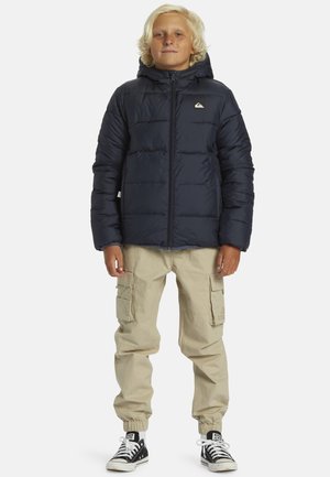 Marinblå puffjacka med huva och quiltat mönster, med en liten logotyp på bröstet; kombinerad med beige cargobyxor och svarta sneakers.