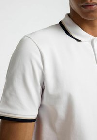 Polo blanc avec une texture unie et un col rayé bleu marine et crème. Manches courtes avec un bord contrastant. Fermeture à boutons.