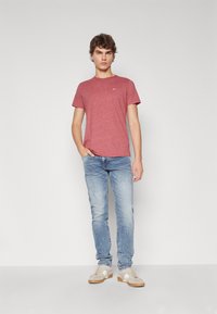 Röd melerad T-shirt med kort ärm, kombinerad med ljusblå slitna jeans och grå sneakers. Enkel logotypdetalj på tröjan.