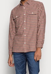 Uomo che indossa una camicia a maniche lunghe a quadri rossi, marroni e bianchi, abbinata a jeans di denim blu, in piedi con le braccia rilassate.