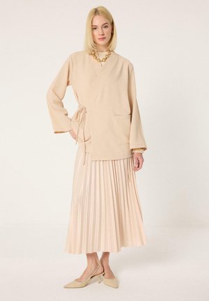 Žena s blond vlasmi, ktorá má na sebe béžový zavinovací kabát, plisovanú midi sukňu, špicaté lodičky so slingback pätou a hrubý zlatý náhrdelník na bielom pozadí.