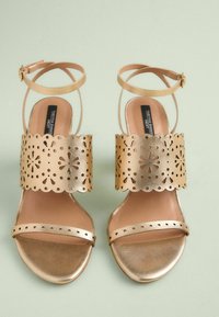Gold heeled sandals with an ankle strap and a perforated wide band. Smooth texture with a glossy finish and cut-out floral details.