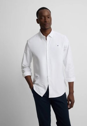 Hombre con una camisa blanca de manga larga con las mangas remangadas y pantalones oscuros, de pie frente a un fondo claro y liso.