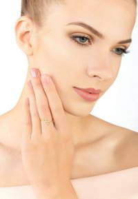 Gold ring with a minimalist design featuring a small clear stone. Model's hand rests on her cheek, showcasing the ring's subtle elegance.