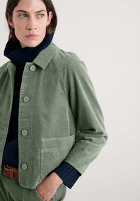 Groene corduroy jas met een kraag, voorzien van knoopsluiting, zijzak en gestructureerde stof, gelaagd over een marineblauwe trui.