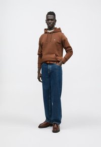 Barna kapucnis pulóver húzózsinórokkal, farmernadrág és barna cipő. A ruha laza szabású és egyszerű dizájnelemeket tartalmaz.