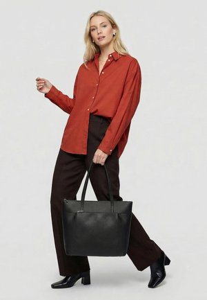 Vrouw in een roestkleurig overhemd met knopen en donkere broek, houdt een grote zwarte leren tote bag vast, staat op één been.