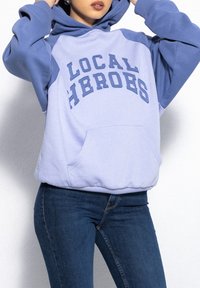 Jasnoniebieska bluza z kapturem z napisem "LOCAL HEROES" w ciemniejszym niebieskim kolorze. Posiada przednią kieszeń, ściągacze na rękawach oraz pojemny kaptur. Noszona z niebieskimi dżinsami.