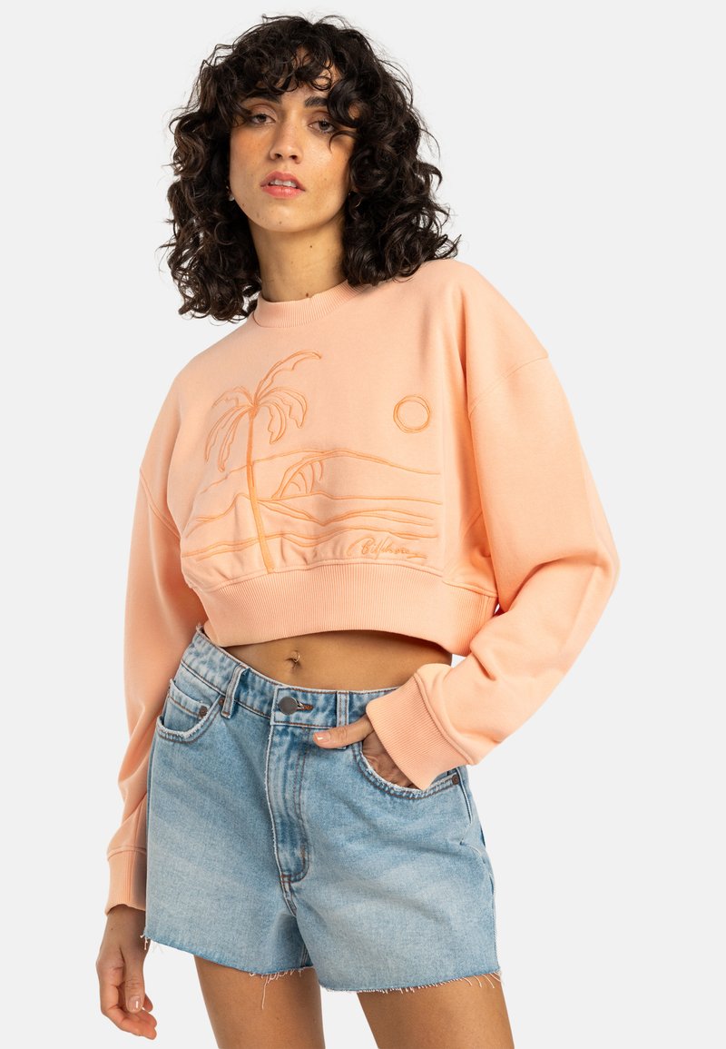 Różowy, krótki sweatshirt w kolorze brzoskwiniowym z haftem przedstawiającym palmę i słońce, w połączeniu z jasnoniebieskimi szortami dżinsowymi z postrzępionym dołem i przednimi kieszeniami.