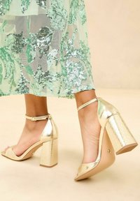 Gold high-heeled sandals with a block heel, featuring an open toe and ankle strap. Textured heel with a woven pattern.