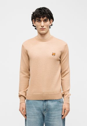 Mladý muž s kučeravými tmavými vlasmi má na sebe béžový sveter s dlhými rukávmi s líščím logom na hrudi a svetlomodré rifle, stojaci pred jednoliatym pozadím.