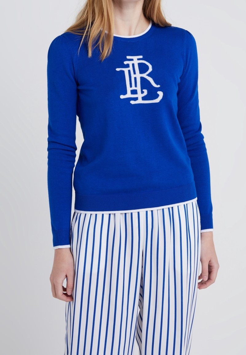 Femme portant un pull bleu à manches longues avec les initiales blanches "TRL" et un pantalon blanc à rayures verticales bleues.