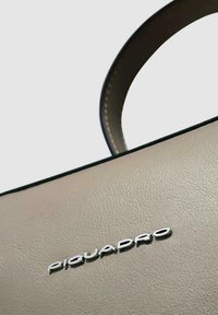Sac à main en cuir beige à texture lisse, garniture foncée et logo "Piquadro" argenté. Présente un design structuré et des poignées arrondies.