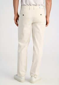 Pantaloni in cotone beige con vestibilità dritta, dotati di tasche posteriori con chiusura a bottone e design contemporaneo. Abbinati a sneaker bianche.