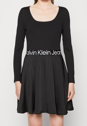 Kobieta ubrana w czarną sukienkę z długim rękawem, dekoltem w serek i rozkloszowanym dołem, z paskiem z logo Calvin Klein Jeans.