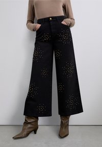 Pantalones anchos negros con aplicaciones de tachuelas doradas y plateadas. Cintura alta, bolsillos delanteros y cierre con botón. Textura de tela lisa.