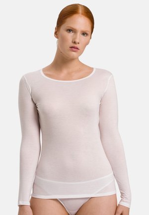 Jeune femme aux cheveux roux portant un haut blanc moulant à manches longues et des sous-vêtements assortis, debout devant un fond clair uni.