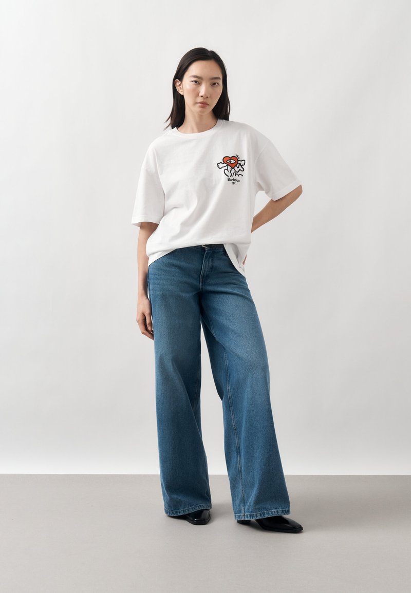 Une femme porte un T-shirt blanc à motif graphique, un jean bleu à jambes larges et des chaussures noires sur un fond de studio uni.