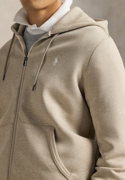 Muž nosiaci béžovú mikinu s kapucňou a zipsom cez bielu košeľu s golierom, ukazujúci predné vrecko mikiny a malé logo na hrudi.