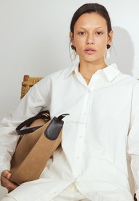 Tan and black handbag with a structured shape and leather accents held by a person in a white button-up shirt. Textured background.