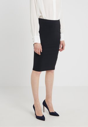 Frau trägt einen schwarzen knieumspielenden Bleistiftrock, eine weiße Bluse mit langen Ärmeln und schwarze High-Heel-Schuhe, steht auf einem schlichten weißen Boden.