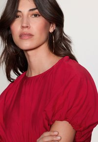 Punainen pusero, jossa on pussihihat ja pyöreä pääntie. Sileä kangaspinta, yhdistetty suuriin kultaisiin renkorenkaisiin korvakoruihin. Hiukset osittain auki.