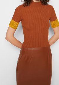 Raitareunruskea toppi lyhyillä hihoilla, jossa on keltaisia neulottuja yksityiskohtia. Yhdistetty ruskeaan nahkahameeseen, joka korostaa tekstuuria ja istuvuutta.